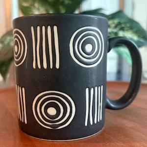 May include: A matte black ceramic mug featuring a repeating pattern of white embossed concentric circles and vertical lines. The mug has a cylindrical body and a curved handle on the right side, with a smooth finish and a consistent dark color.