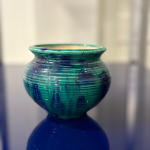 May include: A small, round ceramic pot featuring a vibrant teal and deep blue glaze with a glossy finish. The exterior surface is decorated with horizontal ridges that wrap around the body, leading up to a smooth, flared rim and a wide opening.