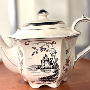 May include: Cream-colored vintage teapot with a black and white landscape scene. The teapot has a silver trim, a dark blue finial, and a curved handle. The landscape features a castle, trees, and water.