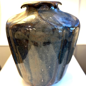 May include: A handcrafted ceramic vase with a unique, fluted design. The vase features a dark, mottled glaze with shades of blue, brown, and gray. The rim has a slightly irregular shape, adding to its artisanal appeal. This decorative vase is suitable for home decor.