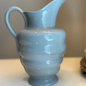 May include: A light blue ceramic pitcher with a glossy finish, featuring a rounded, ribbed body, a curved handle on the side, and a wide pouring spout. The vessel stands on a circular base and has a smooth, reflective surface throughout its exterior.