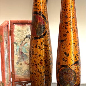 May include: Two tall, slender, cylindrical vases with a glossy orange and black mottled finish. Each vase features a circular medallion with intricate embossed patterns and black accents. Beside them stands a small, folding wooden screen.