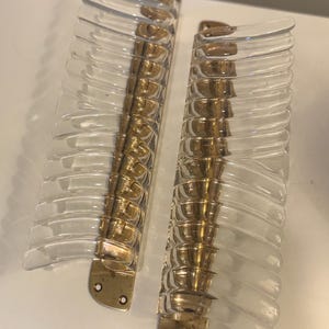 May include: Two vertical wall sconces featuring clear, ribbed glass panels attached to a polished brass metal base. Each base has two circular screw holes at the bottom for mounting. The glass elements are arranged in a series of horizontal fins.