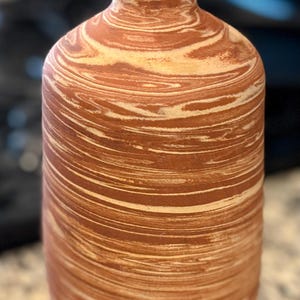 May include: A cylindrical vase with a narrow neck and a flared rim, featuring a marbled pattern of terracotta orange and cream white swirls throughout the exterior surface, creating a continuous horizontal layered effect across the entire body.