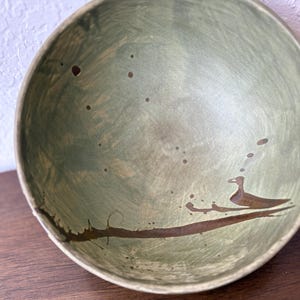 May include: A round ceramic bowl with a sage green glazed interior featuring a hand-painted dark brown branch and a small bird silhouette. The surface has subtle brown speckles and brushstroke textures throughout the interior basin.