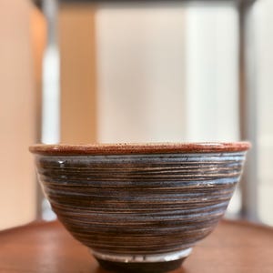 May include: A ceramic bowl featuring a dark brown and blue glazed exterior with horizontal ridged textures. The rim is finished with a speckled reddish-brown glaze, while the base is a solid dark color, creating a distinct contrast in the finish.