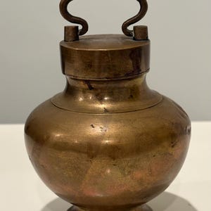 May include: A small, round, antique-style brass vessel with a wide, bulbous body, a narrow neck, and a flat lid featuring a prominent, arched handle. The metal surface displays a weathered, darkened patina with subtle variations in tone and texture.