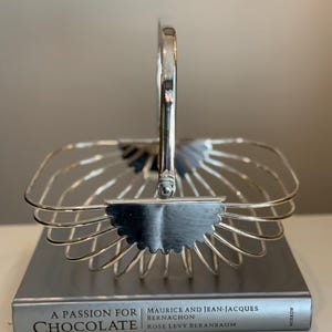 May include: A silver-colored wire basket with a handle, resting on a book titled "A Passion for Chocolate." The basket has a square shape with a scalloped edge and a curved handle. The book's title is visible.