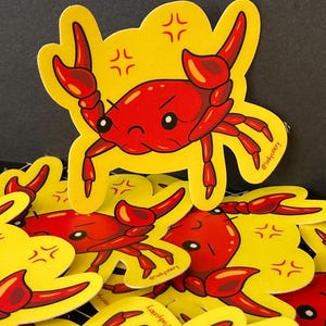 May include: A collection of yellow stickers featuring a cartoon red crab with an angry expression. The crab has large claws and is set against a yellow background with small, stylized angry face symbols. The stickers are scattered.