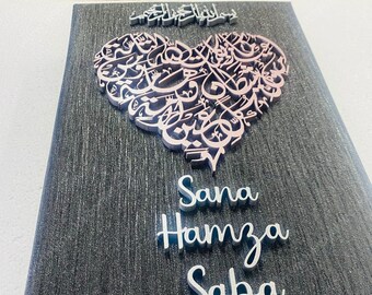 May include: A black canvas with a silver glitter finish features a large heart-shaped design made of intricate Arabic calligraphy. The names "Sana", "Hamza", and "Saba" are written in silver lettering below the heart. The bottom right corner of the canvas has a small logo that reads "The Royal Decors".