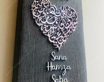 May include: A black canvas with a silver heart-shaped design featuring Arabic calligraphy. The names Sana, Hamza, and Saba are written in silver below the heart.