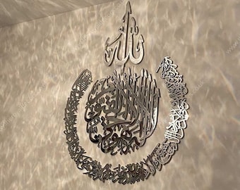 Op de afbeelding: Een zilveren metalen wanddecoratie met Arabische kalligrafie in een cirkelvormig ontwerp. De kalligrafie is in een decoratief lettertype en bevat het woord "Allah" in het midden. De decoratie is bevestigd aan een lichtbeige muur.