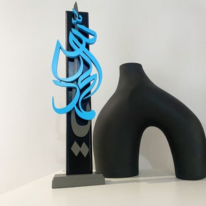 May include: A blue and gray wooden sculpture of Arabic calligraphy on a gray base. The sculpture is next to a black ceramic vase.