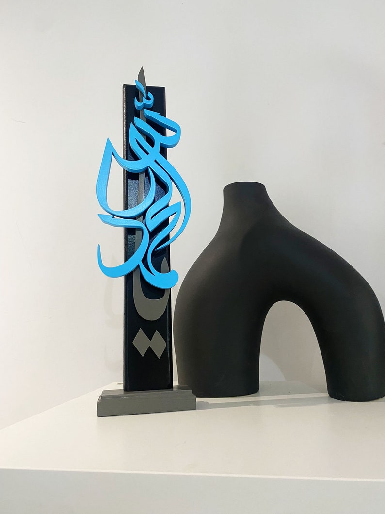 May include: A blue and gray wooden sculpture of Arabic calligraphy on a gray base. The sculpture is next to a black ceramic vase.