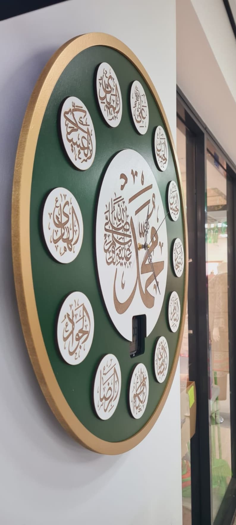 May include: A green and gold circular clock with Arabic calligraphy on a white background. The clock has a gold frame and a white face with black hands. The clock is decorated with 12 smaller circles, each containing Arabic calligraphy.