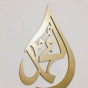 May include: Gold-colored metal wall art in the shape of a teardrop with intricate Arabic calligraphy inside.
