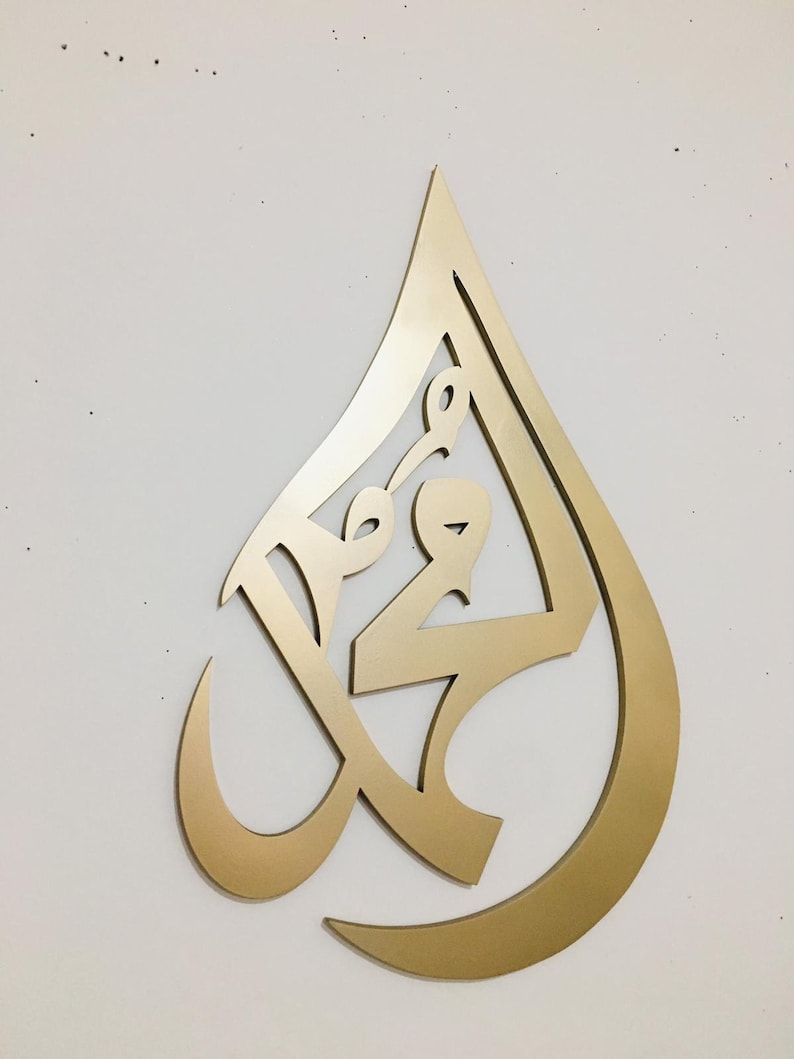 May include: Gold-colored metal wall art in the shape of a teardrop with intricate Arabic calligraphy inside.