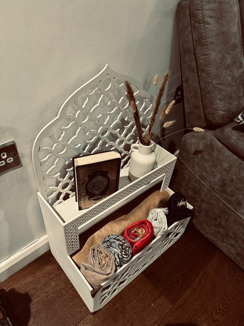 May include: White wooden shelf with intricate cutouts, featuring a book, dried flowers in a vase, and rolled-up fabric. The shelf has two tiers and is designed in a traditional Islamic style.