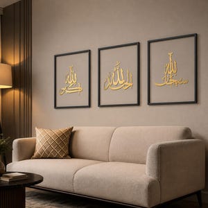 May include: Three framed gold Arabic calligraphy art pieces. Each piece features elegant script on a neutral background, set within black frames. The artwork hangs above a beige sofa with a patterned pillow, creating a warm and inviting living space.