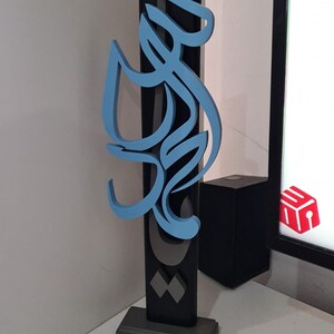 May include: A blue and gray 3D sculpture of Arabic calligraphy on a black base. The sculpture is mounted on a gray base.