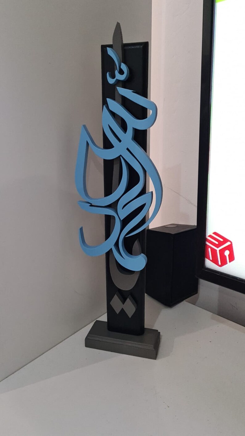 May include: A blue and gray 3D sculpture of Arabic calligraphy on a black base. The sculpture is mounted on a gray base.