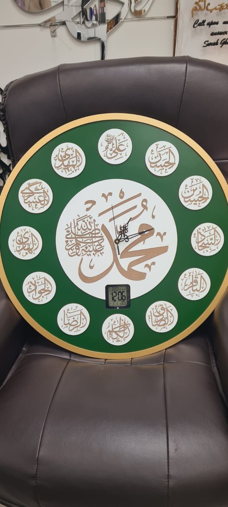 May include: A round, green and gold clock with Arabic calligraphy and a digital display showing the time as 12:06. The clock is on a brown leather chair.