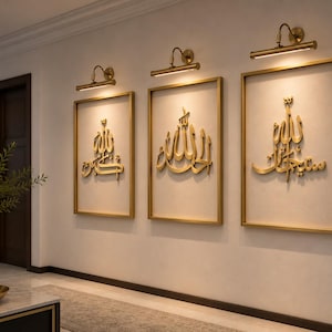 May include: Three framed wall art pieces with gold Arabic calligraphy. Each piece is rectangular with a gold frame and a cream-colored background. Above each piece is a gold-toned adjustable picture light. The wall is a light beige color.