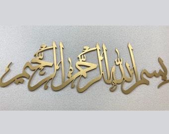 May include: Gold-colored Arabic calligraphy wall art. The script is a flowing design, likely a religious phrase. The artwork is mounted on a textured, off-white surface, creating a striking contrast. The piece is suitable for home decor.