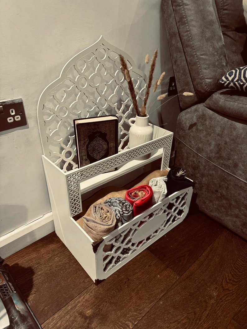 May include: White wooden shelf with intricate cutouts in the shape of an arch. The shelf has two tiers and is filled with items like a book, a vase with dried flowers, and rolled-up fabric.