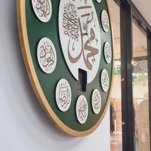 May include: A round, green and gold clock with Arabic calligraphy. The clock has a white center with a large Arabic inscription and smaller inscriptions around the edge. The clock is hanging on a white wall.