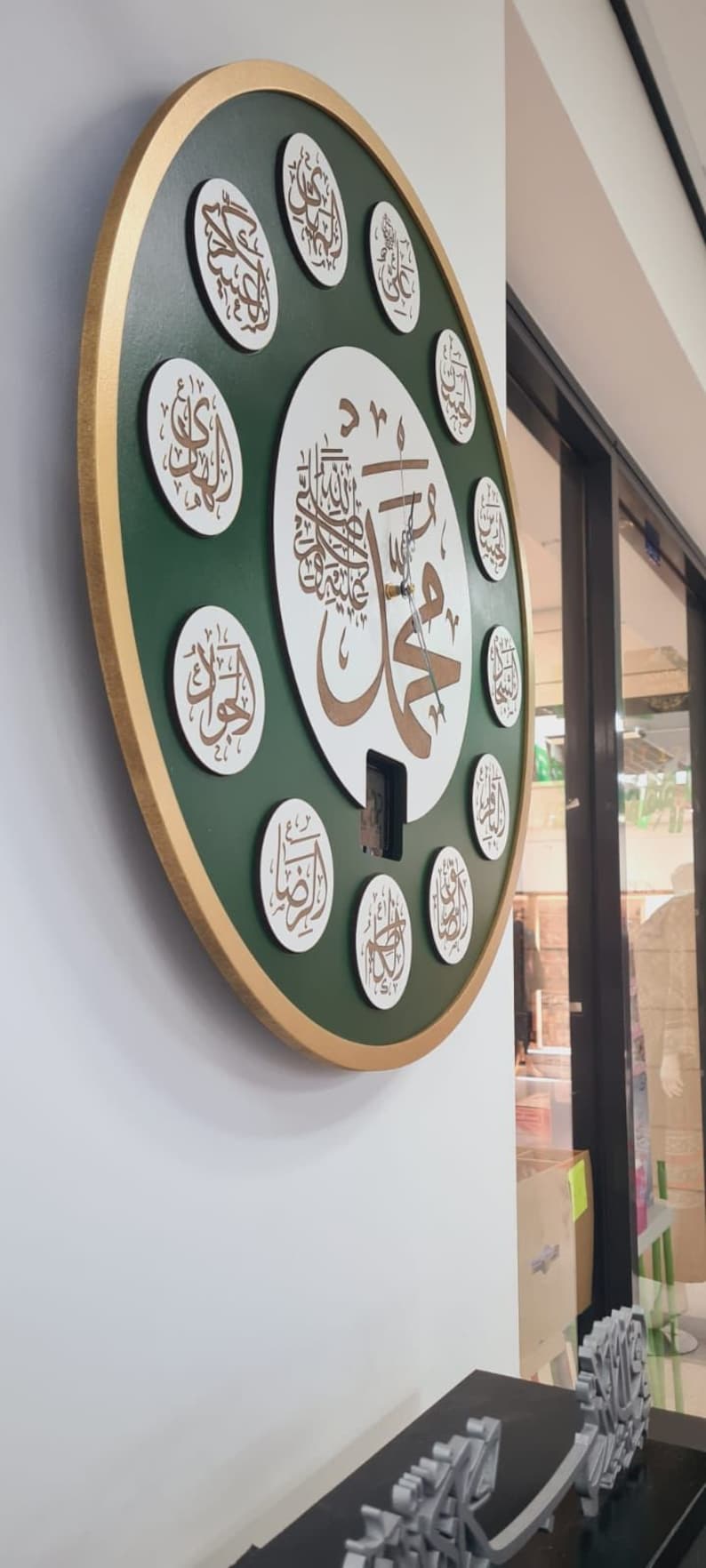 May include: A round, green and gold clock with Arabic calligraphy. The clock has a white center with a large Arabic inscription and smaller inscriptions around the edge. The clock is hanging on a white wall.