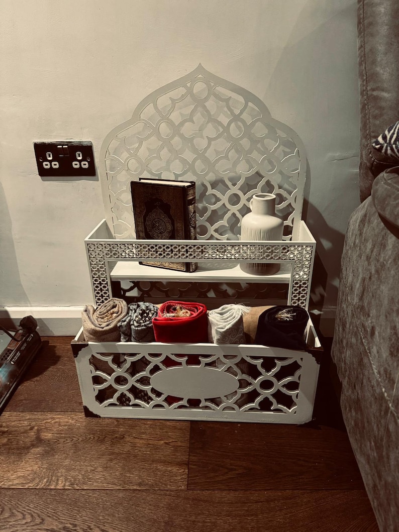 May include: White wooden shelf unit with intricate cut-out designs. The shelf has two tiers and a storage basket below. The top tier holds a book and a white vase. The bottom tier holds several rolled-up fabric items.