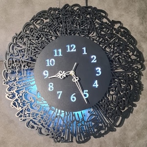 May include: A black clock with Arabic calligraphy around the edge. The clock face is black with white numbers and silver hands. The clock is illuminated from behind with a blue light.