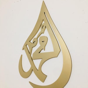 May include: Gold-colored metal wall art in the shape of a teardrop with intricate Arabic calligraphy inside.