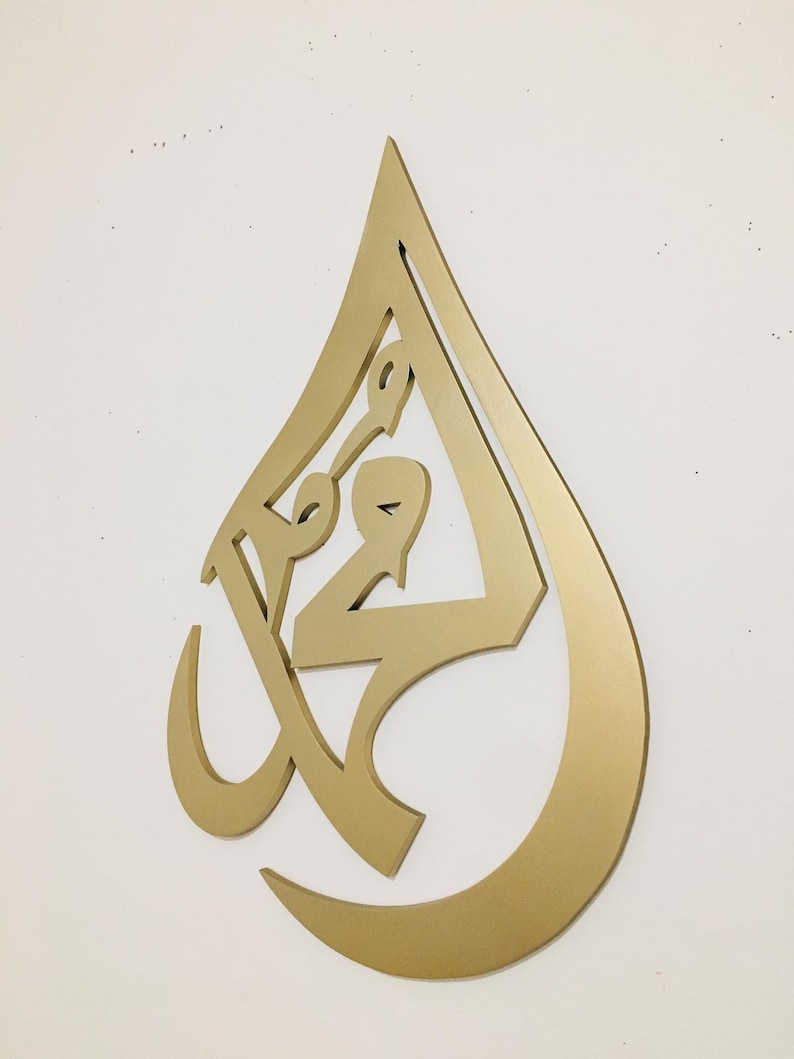 May include: Gold-colored metal wall art in the shape of a teardrop with intricate Arabic calligraphy inside.