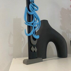 May include: A blue and gray wooden sculpture with Arabic calligraphy on a gray base. The sculpture is on a white shelf.