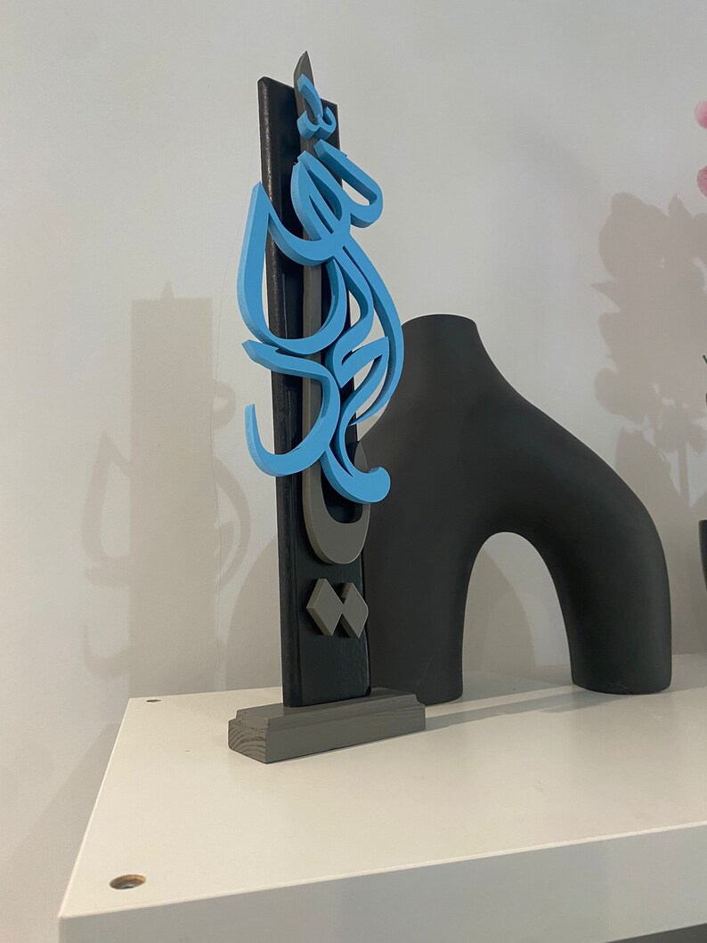 May include: A blue and gray wooden sculpture with Arabic calligraphy on a gray base. The sculpture is on a white shelf.