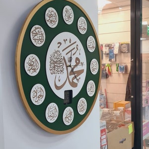 May include: A green and gold circular wall clock with Arabic calligraphy and a digital display. The clock is surrounded by 12 smaller circles, each containing Arabic calligraphy. The clock is hanging on a white wall.