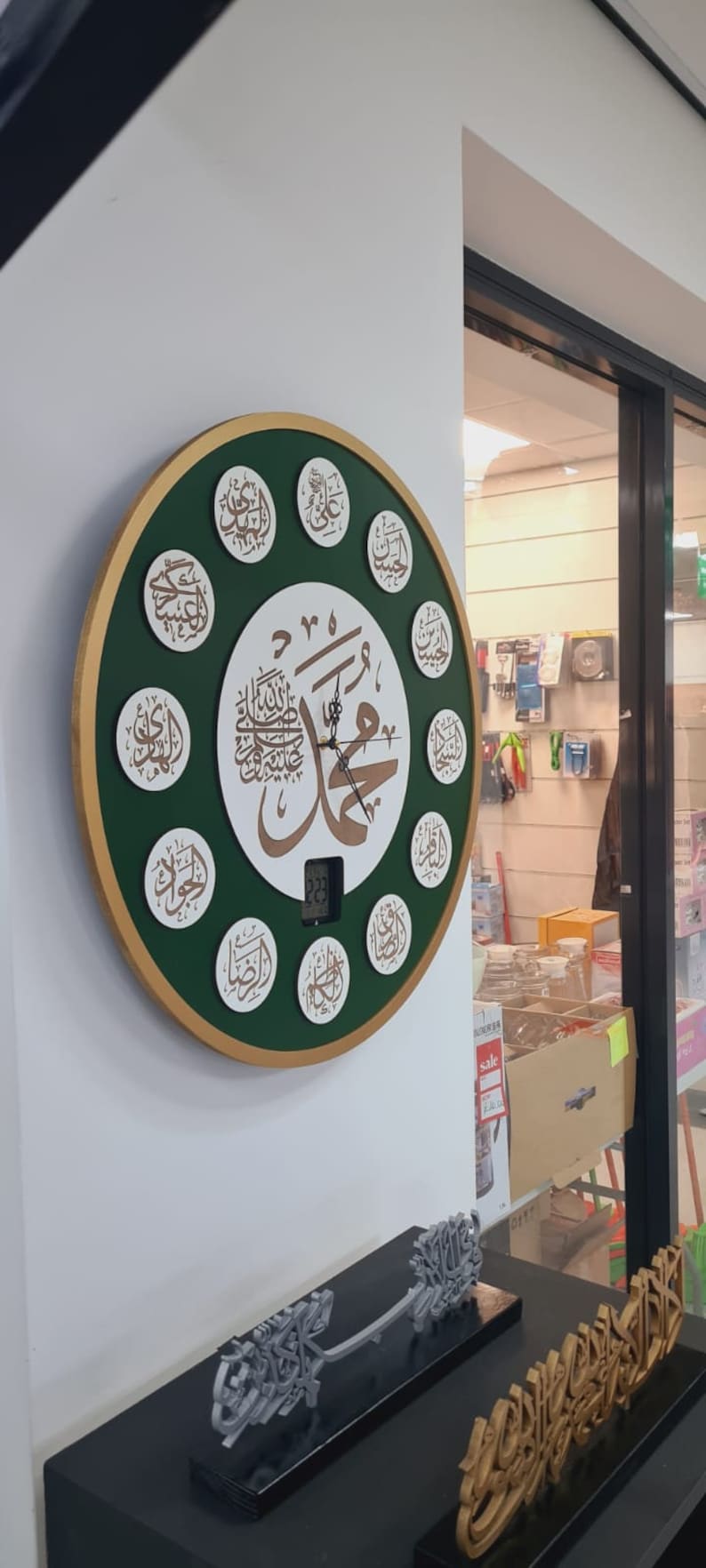 May include: A green and gold circular wall clock with Arabic calligraphy and a digital display. The clock is surrounded by 12 smaller circles, each containing Arabic calligraphy. The clock is hanging on a white wall.