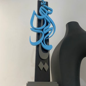 May include: A blue and gray wooden sculpture with Arabic calligraphy. The sculpture is mounted on a gray base and stands on a white surface.