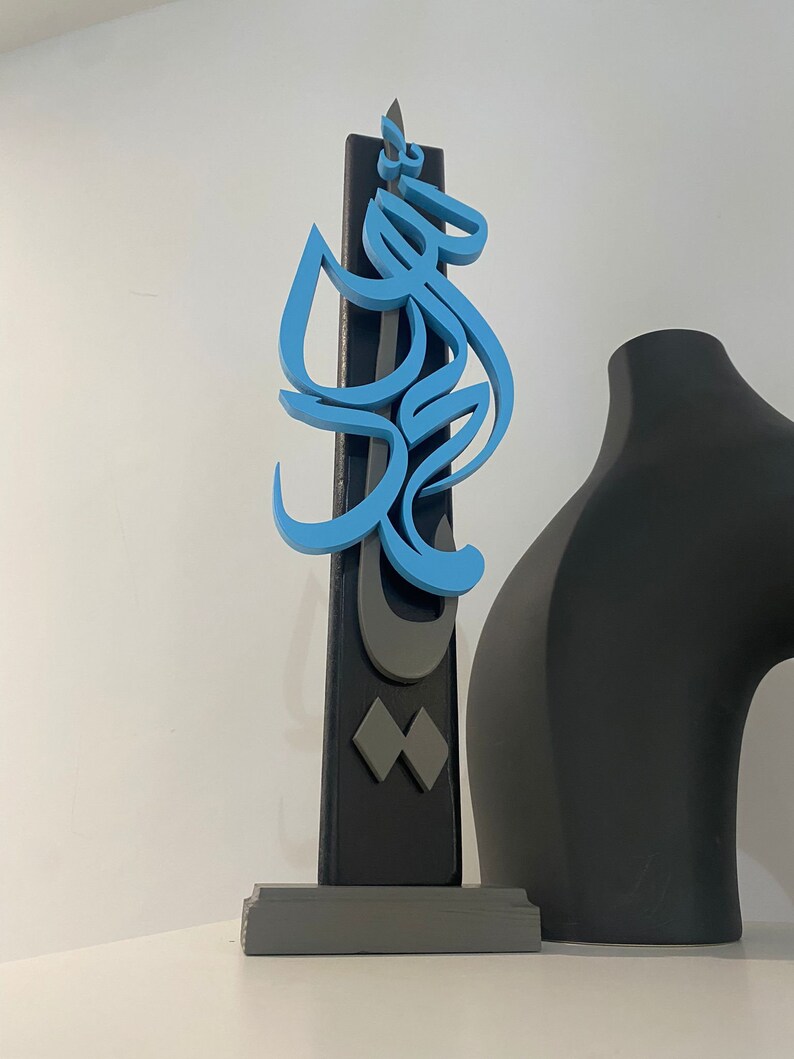 May include: A blue and gray wooden sculpture with Arabic calligraphy. The sculpture is mounted on a gray base and stands on a white surface.