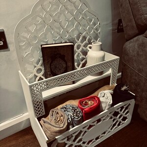 May include: White wooden shelf with intricate cutouts in the shape of an arch. The shelf has two tiers and is decorated with a white vase and a book. The bottom tier has a storage compartment with rolled-up fabric.