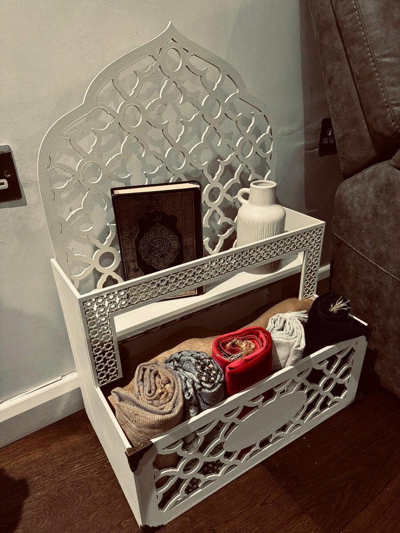 May include: White wooden shelf with intricate cutouts in the shape of an arch. The shelf has two tiers and is decorated with a white vase and a book. The bottom tier has a storage compartment with rolled-up fabric.