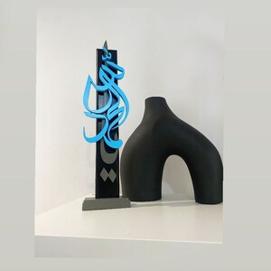 May include: A black and blue wooden sculpture with Arabic calligraphy on a gray base, next to a black ceramic vase with a curved shape.