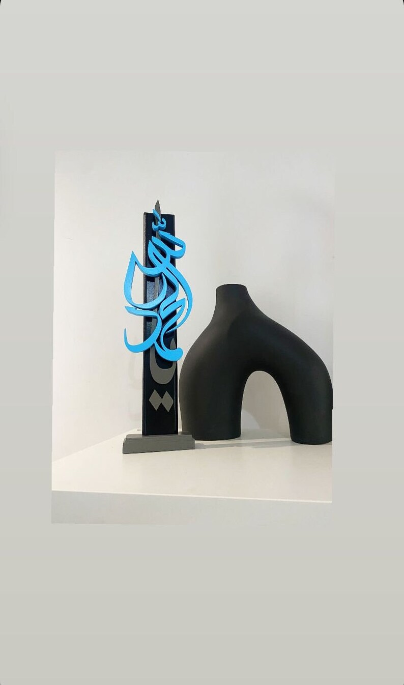 May include: A black and blue wooden sculpture with Arabic calligraphy on a gray base, next to a black ceramic vase with a curved shape.