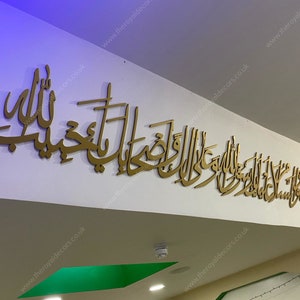 Op de afbeelding: Gouden Arabische kalligrafie wandkunst met de tekst "Allah" en andere Arabische woorden. De kunst is gemonteerd op een witte muur.