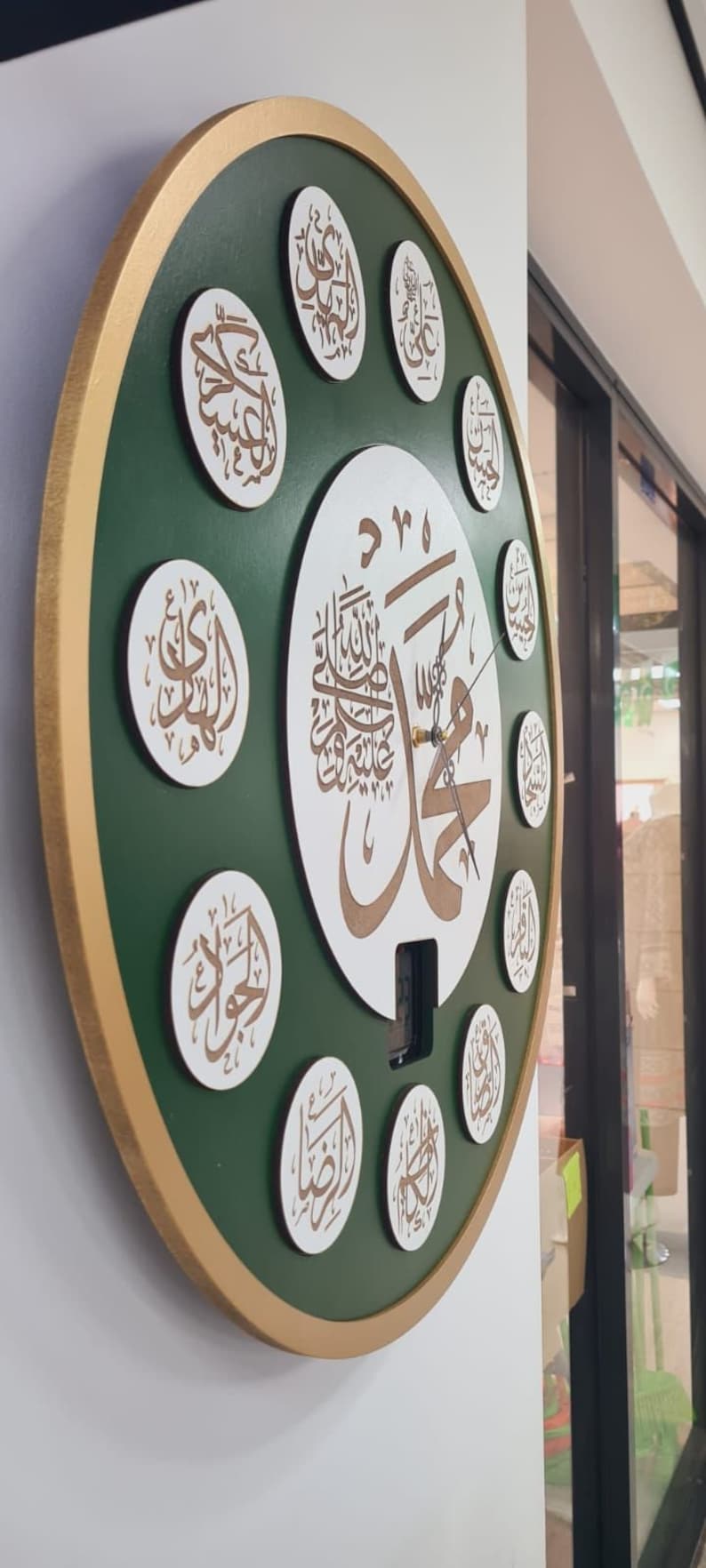 May include: A green and gold circular wall clock with Arabic calligraphy in white and gold. The clock has a white center with a large Arabic inscription and a smaller clock face with hands.