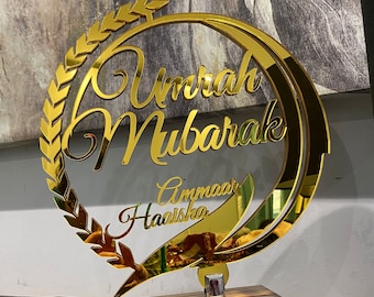 Peut inclure: Plaque en acrylique miroir doré avec le texte "Umrah Mubarak" et les noms "Ammaar" et "Haaisha" dans une police décorative. La plaque est montée sur une base en bois.