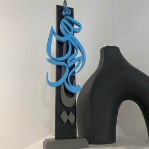 May include: A blue and gray wooden sculpture with Arabic calligraphy on a black background. The sculpture is on a gray base and is next to a black vase.