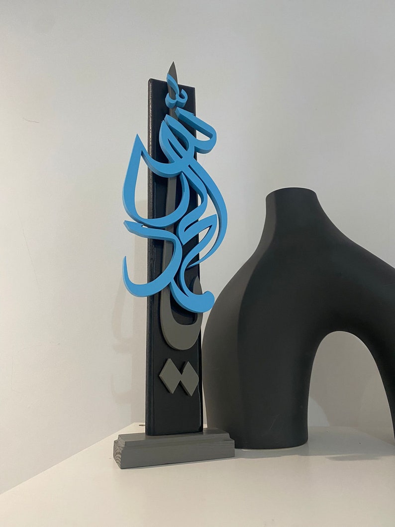 May include: A blue and gray wooden sculpture with Arabic calligraphy on a black background. The sculpture is on a gray base and is next to a black vase.